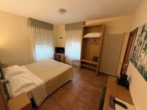 A hotel room with a bed, a desk, a shelf with hanging clothes, a radiator, and a painting on the wall. - Hotel Trieste (Tarvisio)