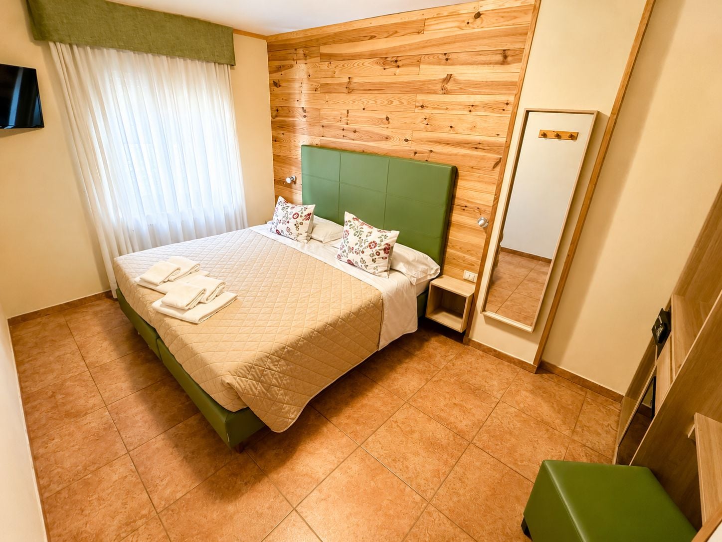 A hotel room with a bed, a green headboard, a bedside table, a mirror, and a green bench.