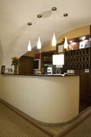 Uma área de recepção de hotel com balcão curvo, um vaso de flores, uma mesa com computador e abajur, e um suporte de chaves fixado na parede.