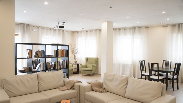 A modern living room with a beige sofa, a coffee table, a bookshelf, a green armchair, and a dining area with a table and chairs. - Hotel Trieste (Tarvisio)