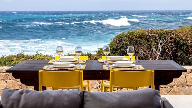 A dining table set for two with white plates and wine glasses, overlooking the ocean.