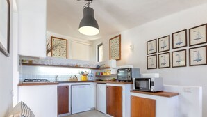 A compact kitchen with white cabinets, a built-in microwave, a toaster, and a coffee maker. There are framed pictures on the wall and a wooden bench in the corner.