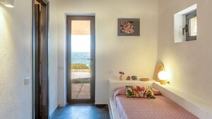 A bedroom with a bed, a door leading to a balcony, a small shelf with decorative items, and a wall-mounted light.