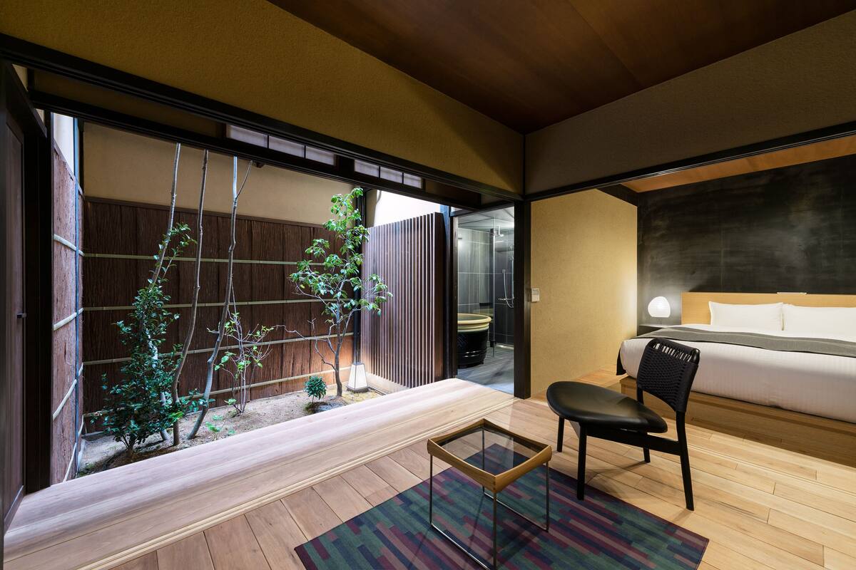 Une chambre d’hôtel moderne avec un plancher en bois, un lit recouvert de draps blancs, une chaise noire, une petite table et une vue sur un jardin, accessible par une porte coulissante.