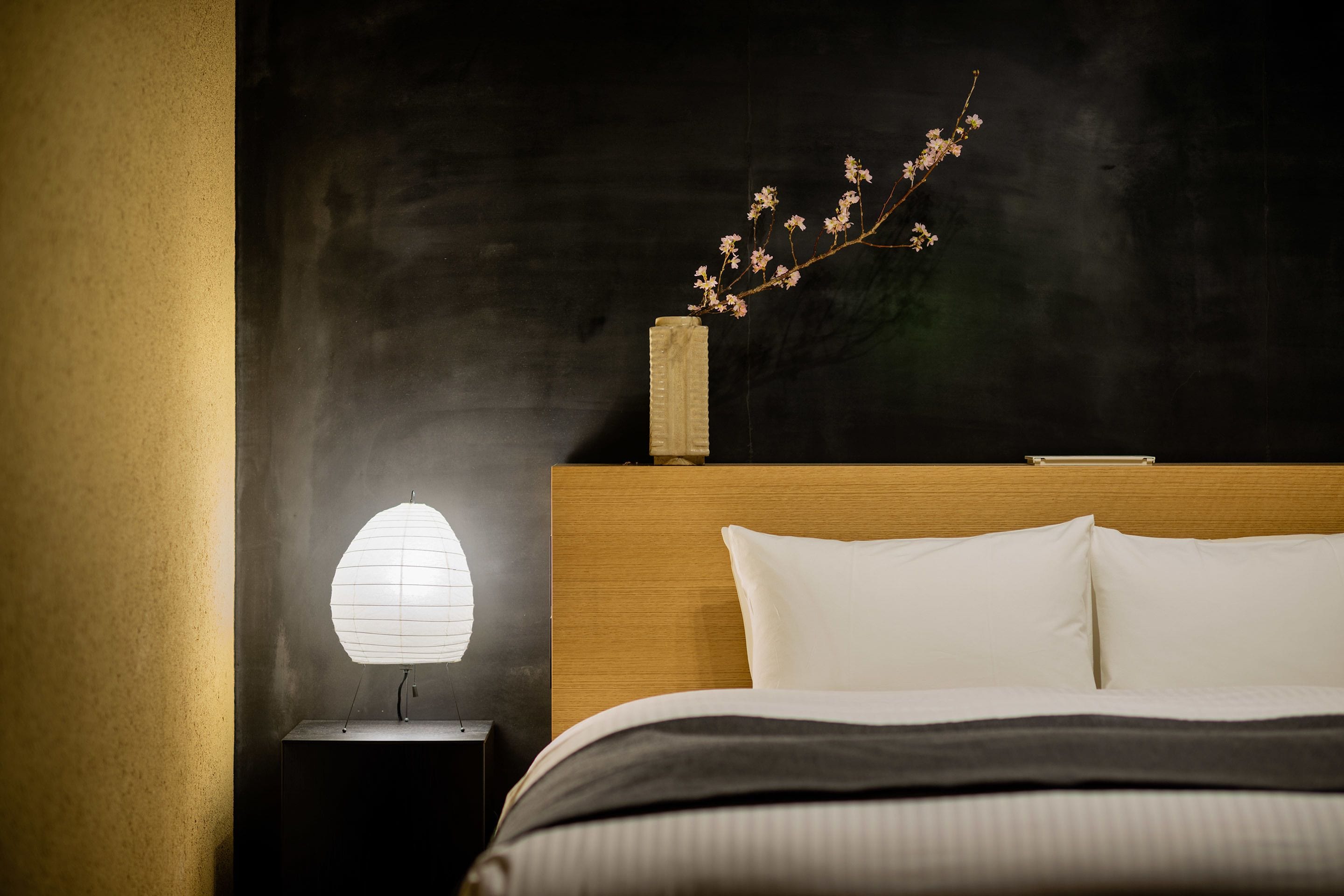A neatly made bed with white linens, a bedside lamp, and a vase with a branch of flowers on a wooden headboard.