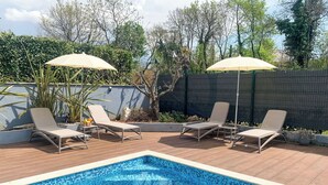 Une piscine entourée d’une terrasse en bois et d’un petit muret, avec des chaises longues et des parasols à proximité.