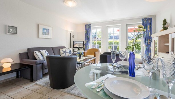 Ein modernes Wohnzimmer mit einer Couch, einem Esstisch für zwei Personen und einem großen Fenster mit Vorhängen.