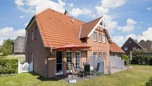 Ein zweistöckiges Backsteinhaus mit rotem Ziegeldach, einer überdachten Terrasse mit Gartenmöbeln und einem weißen Zaun.