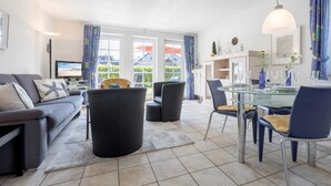 Ein modernes Wohnzimmer mit einer Couch, einem Glastisch und Stühlen. Links befindet sich ein Fernseher und ein Fenster mit Vorhängen.