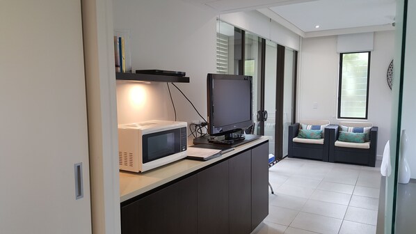 A modern kitchen with a microwave, a flat-screen TV, and a glass door leading to a seating area with a cushioned bench.