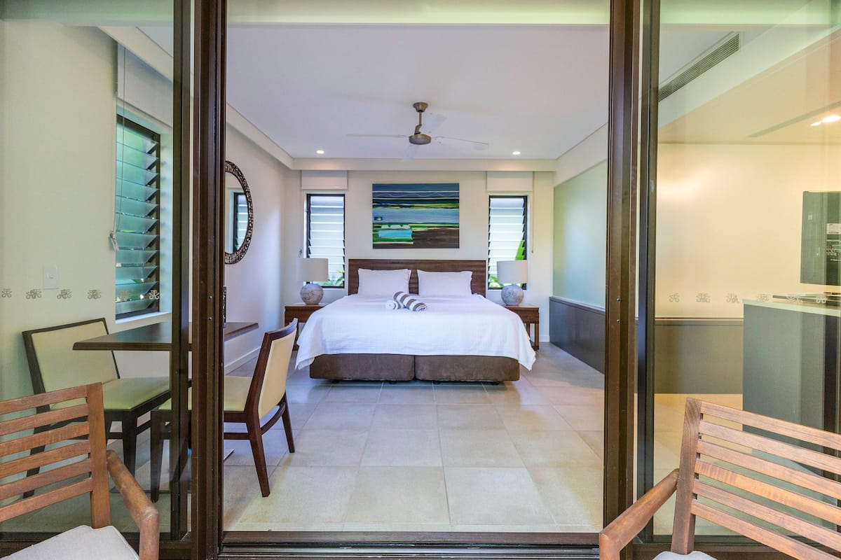 Quarto moderno com uma cama grande, duas mesas de cabeceira, um ventilador de teto e vista para o exterior pelas janelas.