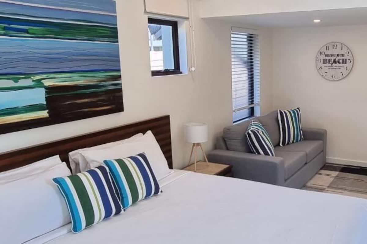 Quarto de hotel com uma cama, um sofá cinza, um relógio na parede e vista para a praia.