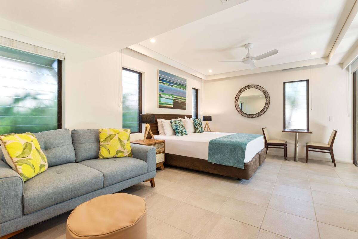 Quarto moderno com sofá, cama e cadeiras, com grandes janelas e ventilador de teto.
