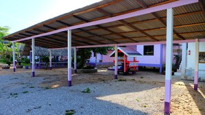 A covered outdoor area with a gravel floor, a red bench, and a building with a purple and white facade in the background. - Rungarun Resort (Chaiya)