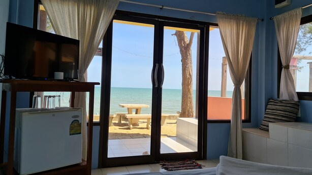 A room with a bed, a TV on a wooden stand, a small refrigerator, and a view of the beach through a glass door with curtains. - Rungarun Resort (Chaiya)