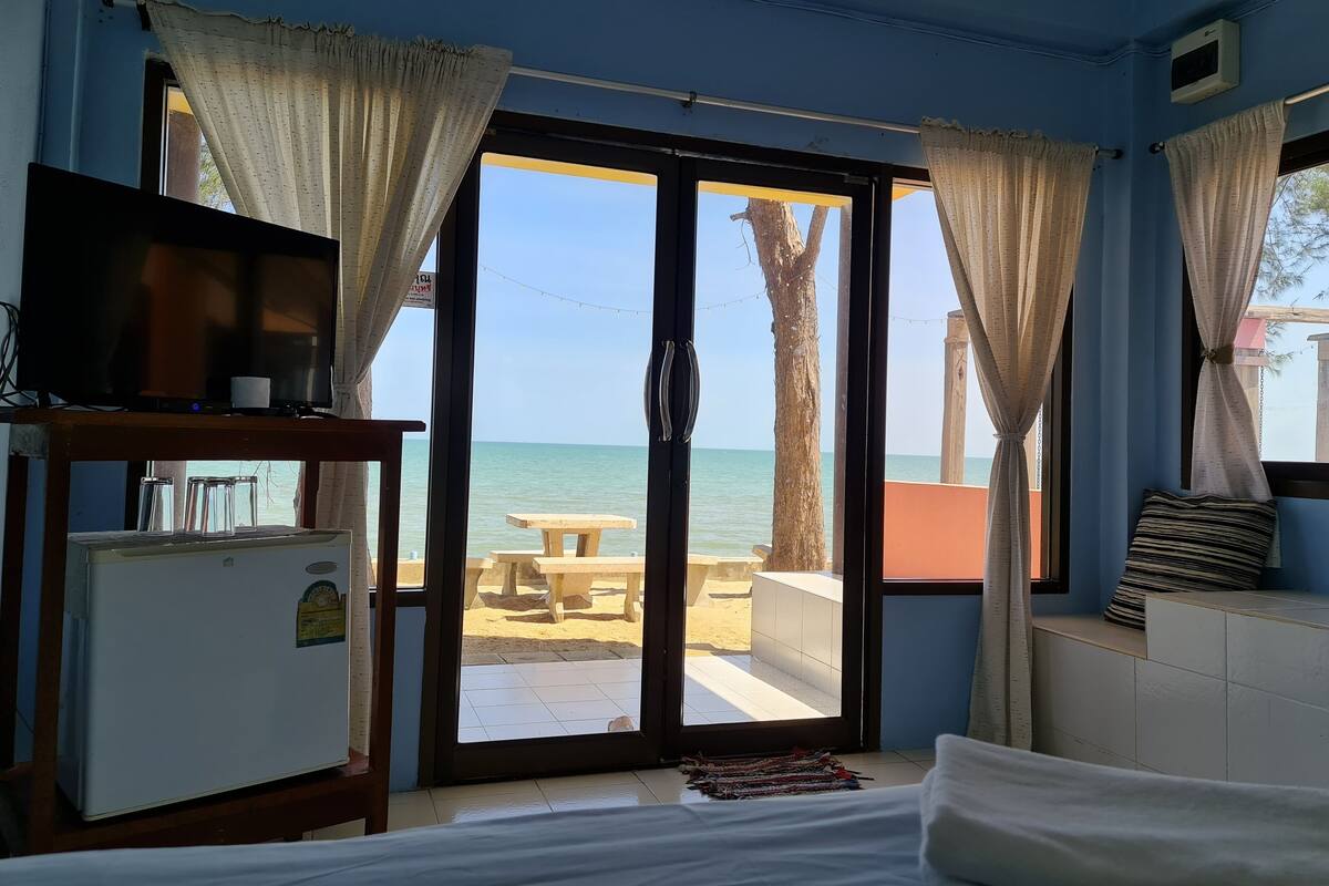 A room with a bed, a TV on a wooden stand, a small refrigerator, and a view of the beach through a glass door with curtains.