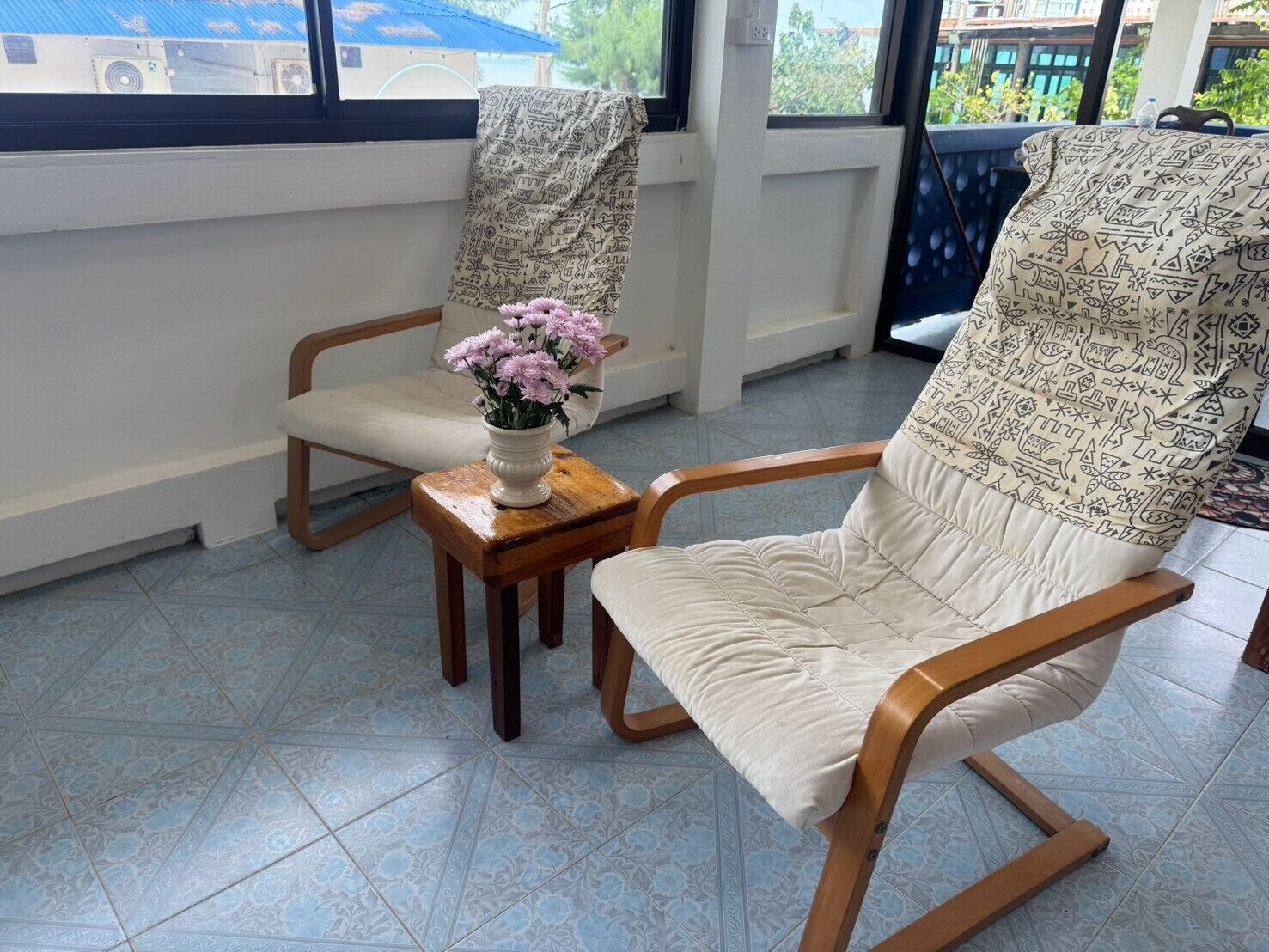 Una habitación con un ventanal grande, una silla con un cojín estampado, una pequeña mesa de madera con un florero y vistas a edificios.