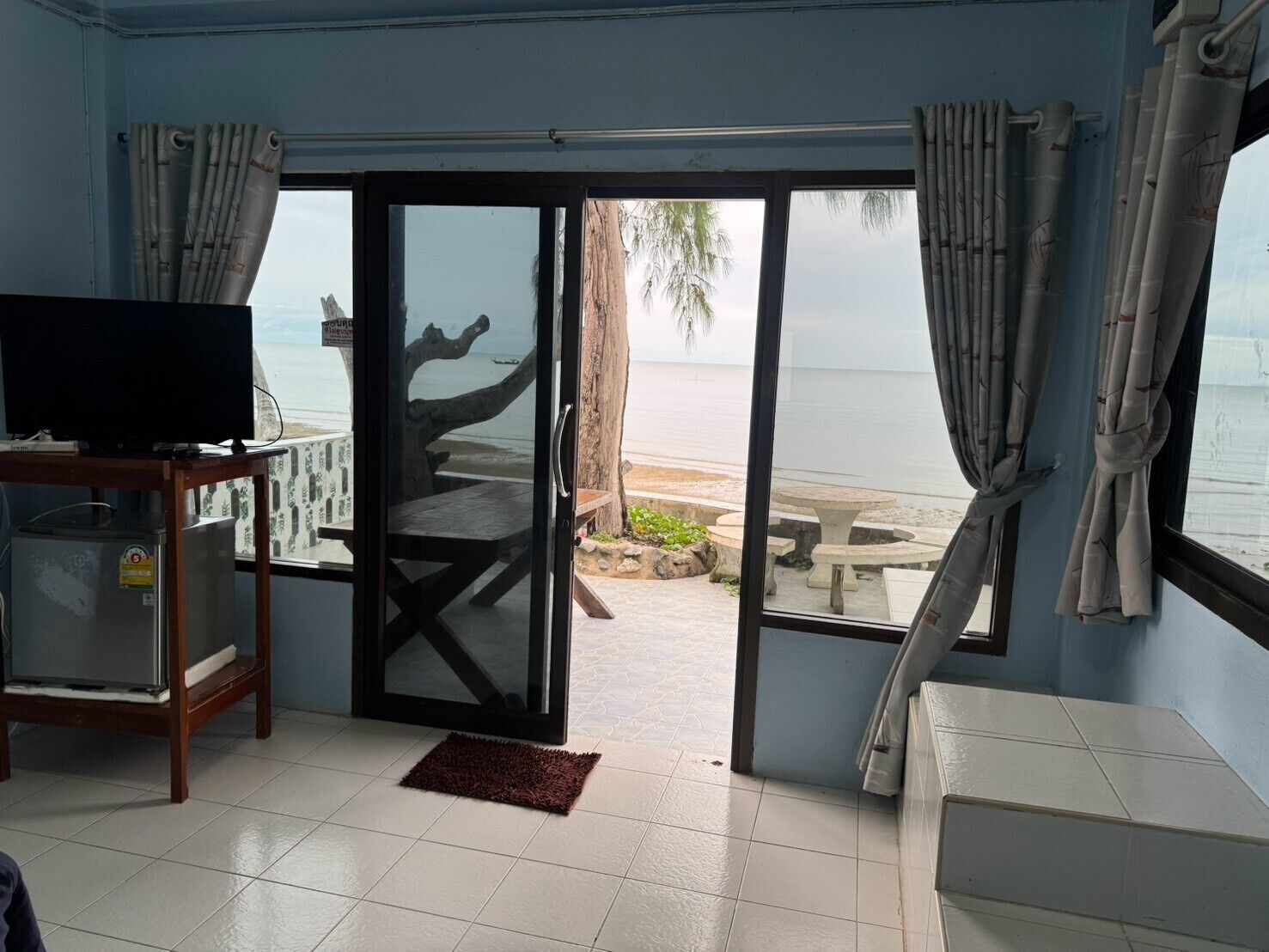 A room with a television on a stand, a sliding glass door leading to a beach, and a small table with chairs outside.