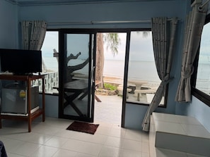 A room with a television on a stand, a sliding glass door leading to a beach, and a small table with chairs outside. - Rungarun Resort (Chaiya)