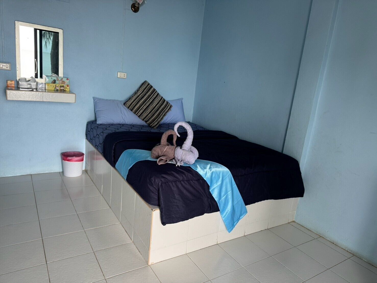 A single bed with a blue and black bedspread, a striped pillow, and a towel shaped like a swan. A small trash bin is visible to the left.