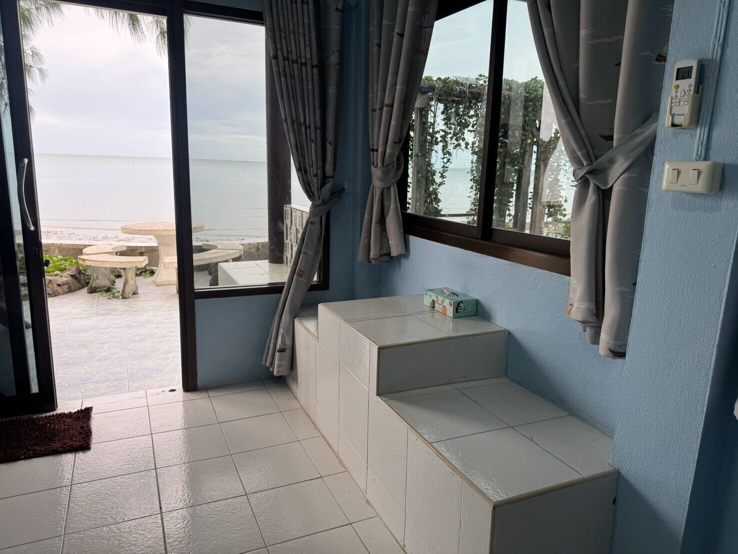 A room with a view of the beach, a small table, a bench, and a thermostat on the wall.
