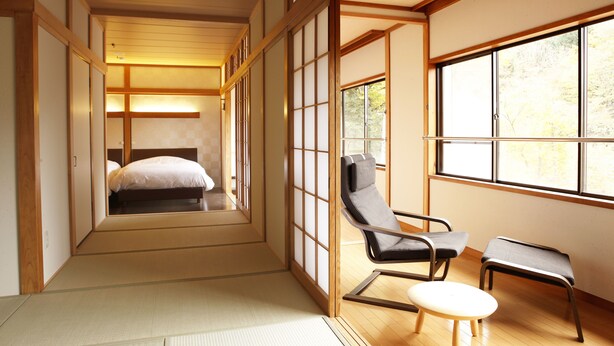 A traditional Japanese room with tatami flooring, a low bed, a wooden sliding door, a window with a view of trees, and a seating area with a chair and stool. - Hakone Fura (Hakone)