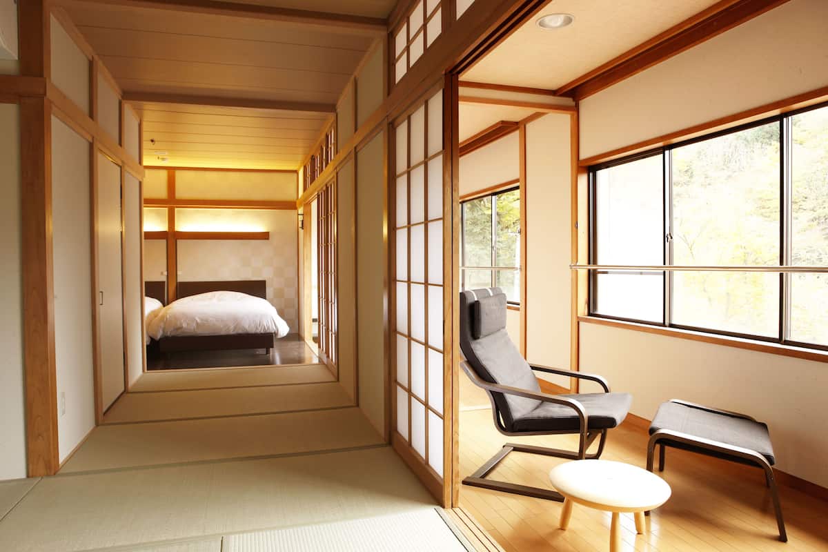 Una habitación japonesa tradicional con suelo de tatami, una cama baja, una puerta corrediza de madera, una ventana con vistas a árboles y un rincón de descanso con una silla y un taburete.
