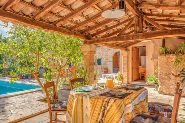 Un área de comedor al aire libre techada, con piscina, muros de piedra y plantas en macetas.