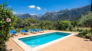 Piscina com cadeiras de descanso azuis, cercada por vegetação e montanhas ao fundo.