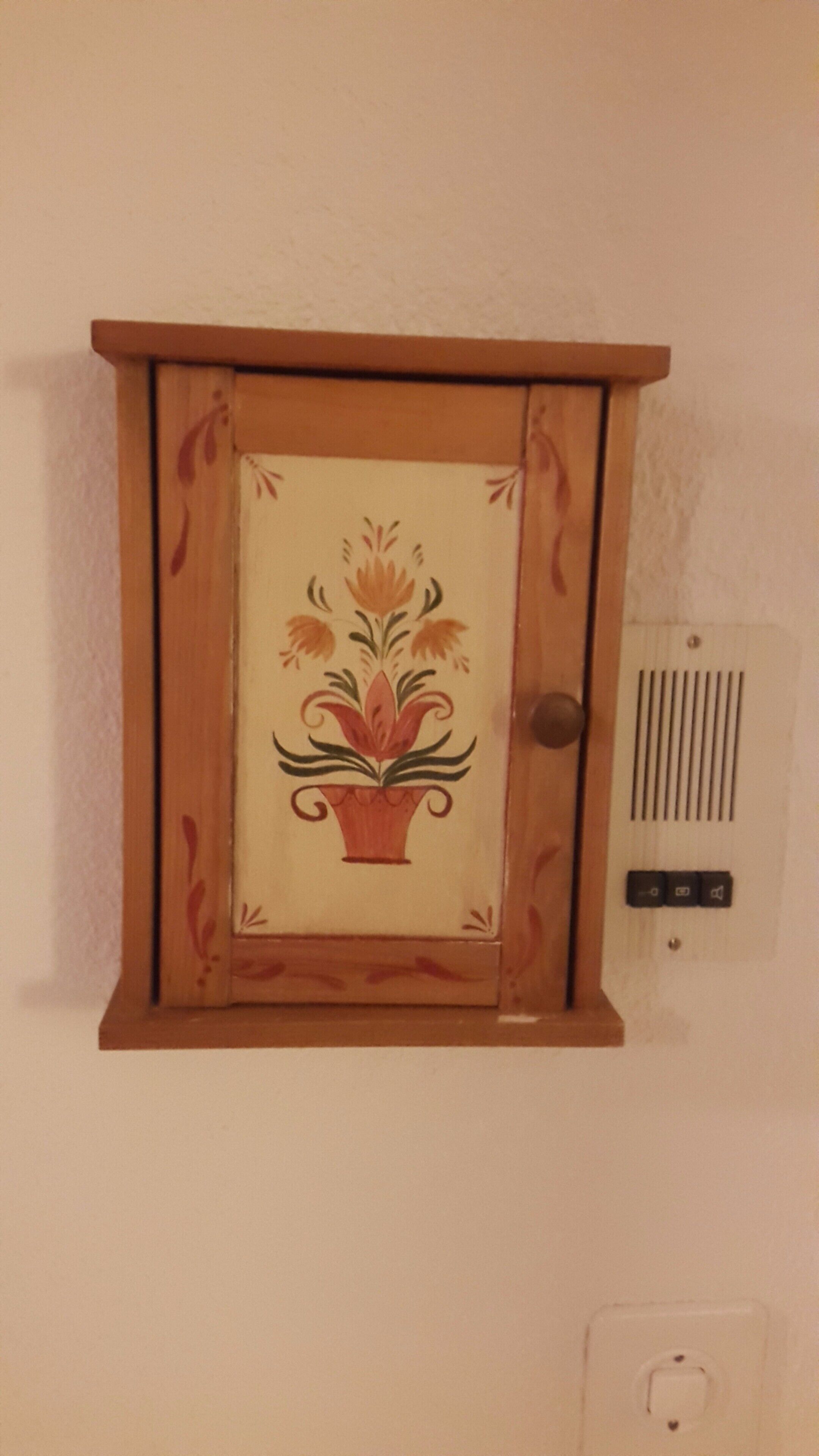 Un armadietto in legno con decorazioni floreali, uno sportello e un termostato fissato alla parete.