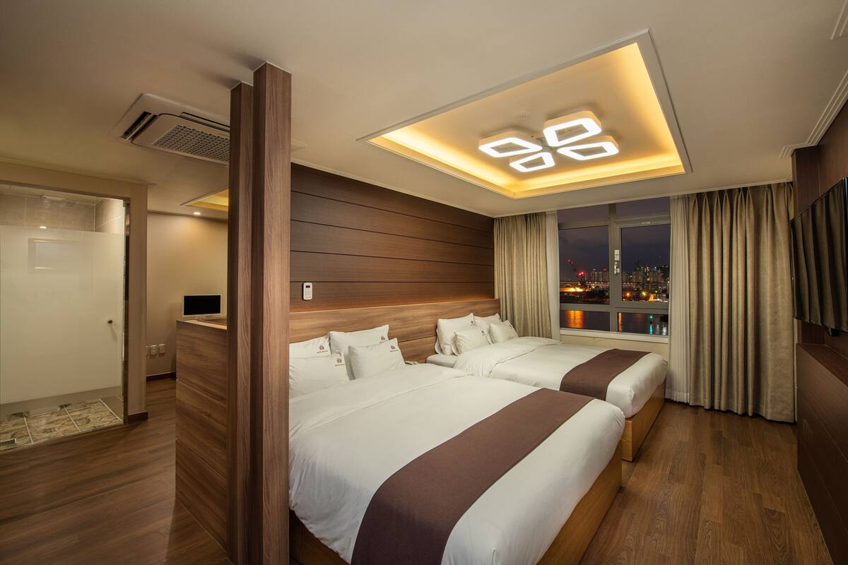 A hotel room with two beds, a city view, a ceiling light fixture, and a wooden floor.