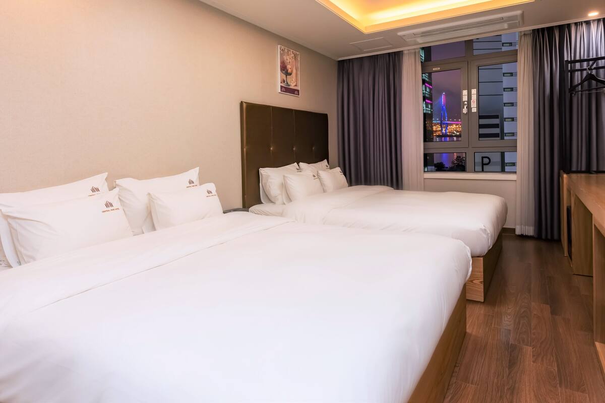 A hotel room with two beds, a wooden floor, a window with curtains, and a view of a cityscape at night.