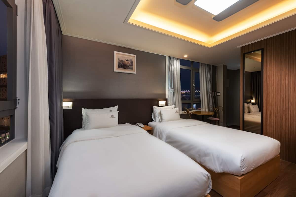 A hotel room with two beds, a large mirror, a window with curtains, and a city view.