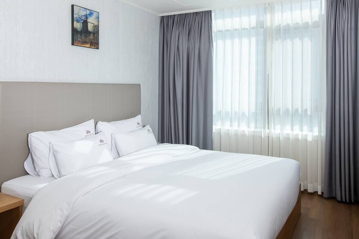A neatly made bed with white linens and a headboard, a bedside table, and a window with sheer curtains and a gray drape.