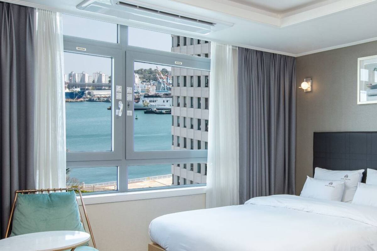 A hotel room with a bed, a chair, a window with a view of a harbor and buildings, and a framed picture on the wall.