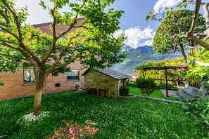 Une maison en pierre avec un jardin, un arbre et une montagne en arrière-plan.