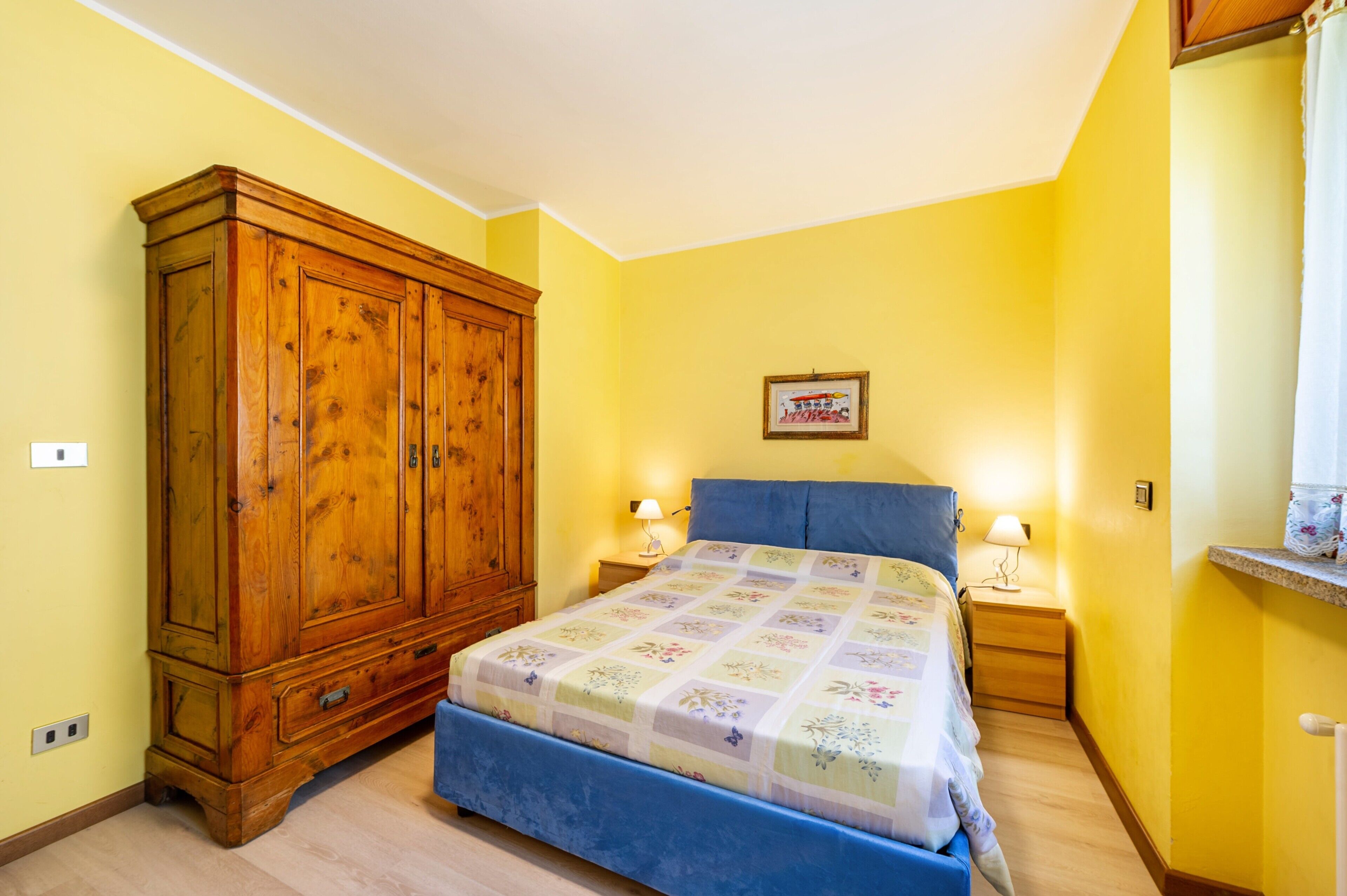 Ein Schlafzimmer mit einem großen Holzschrank, einem Bett mit floralem Überwurf, zwei Nachttischlampen und einem gerahmten Bild an der Wand.