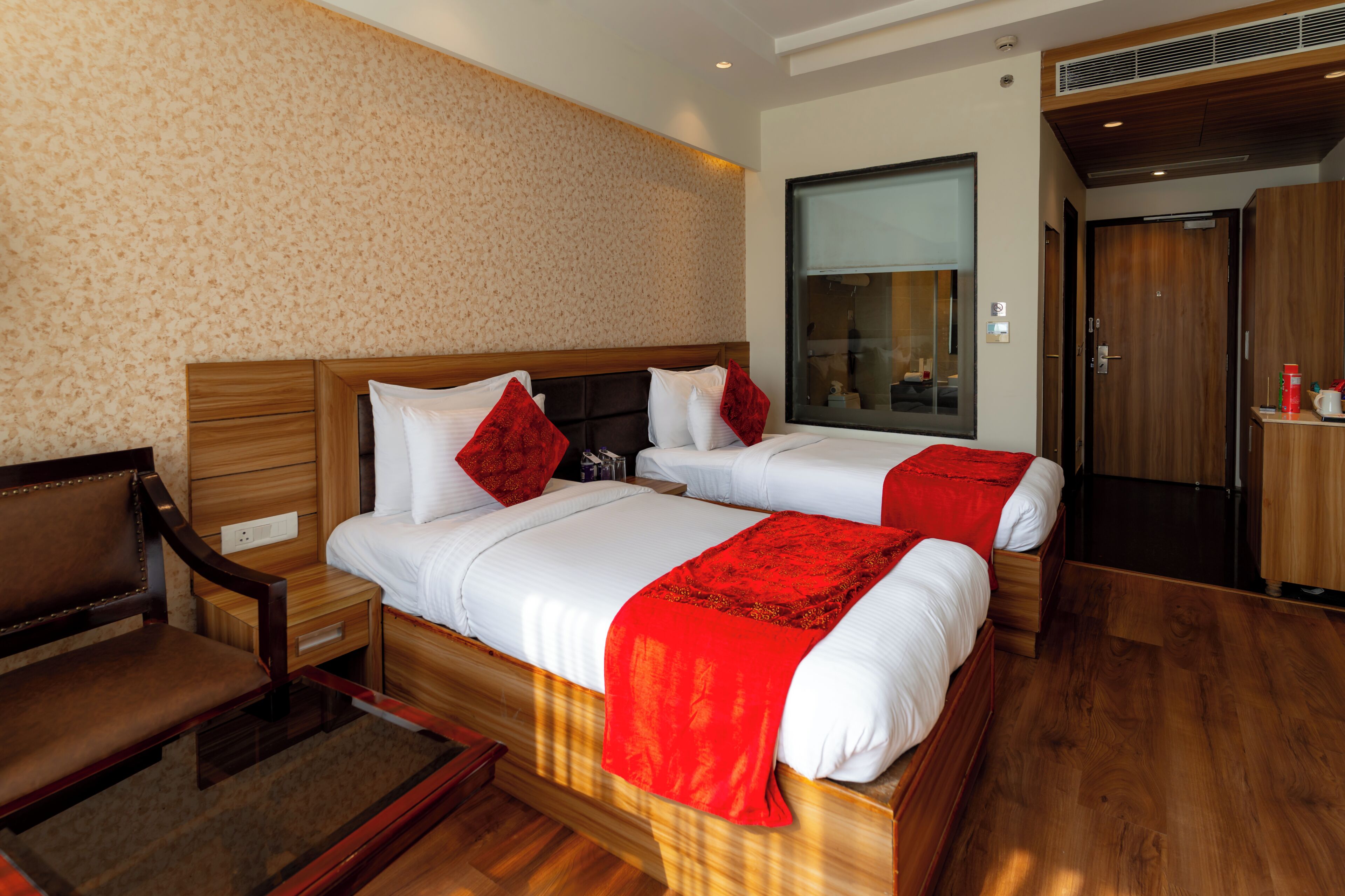 Una habitación de hotel con dos camas, cabeceros de madera, un banco de madera, una mesita de noche y un ventanal grande.