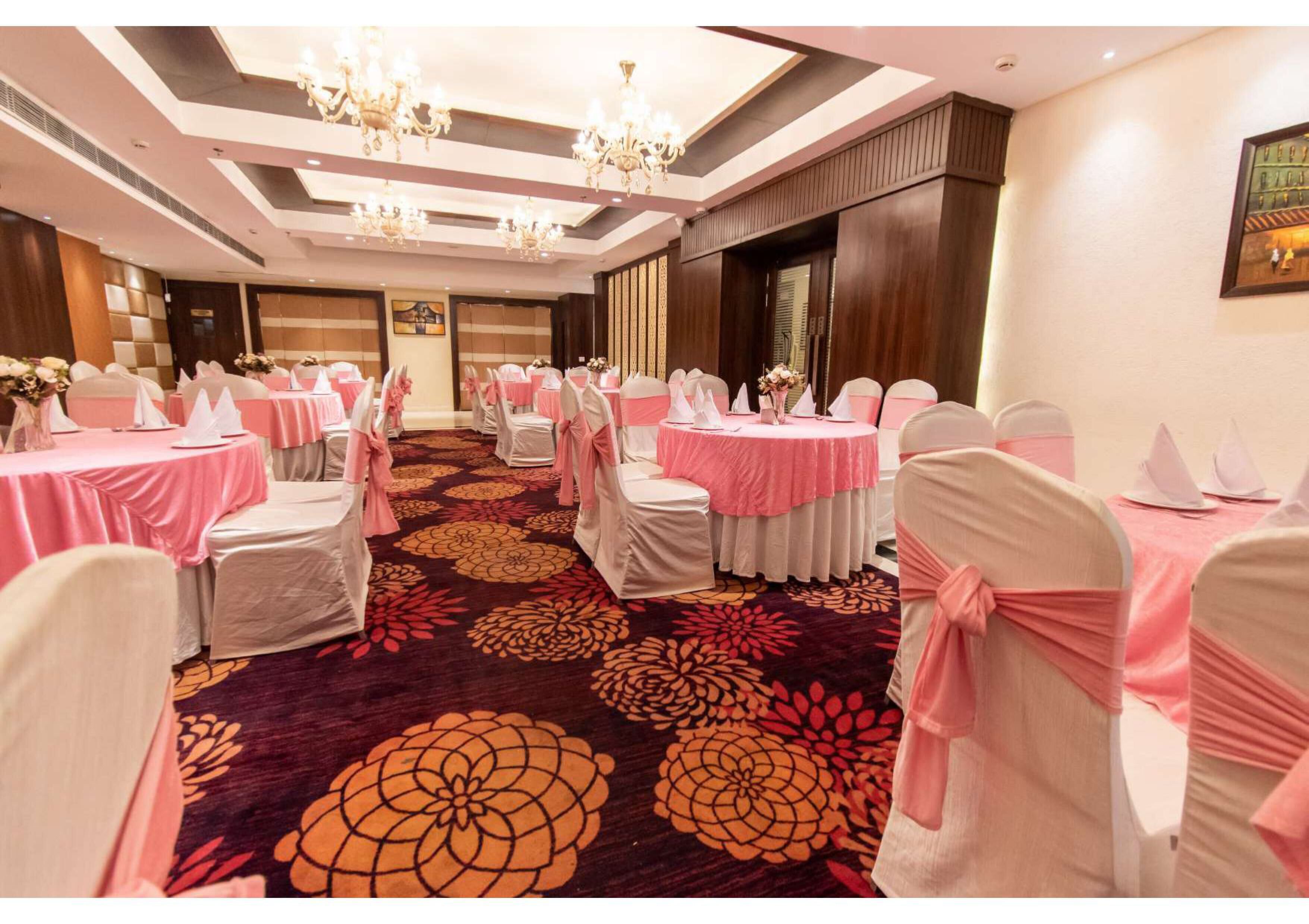 Un salón de banquetes con mesas redondas, sillas con cintas rosas y una alfombra floral.