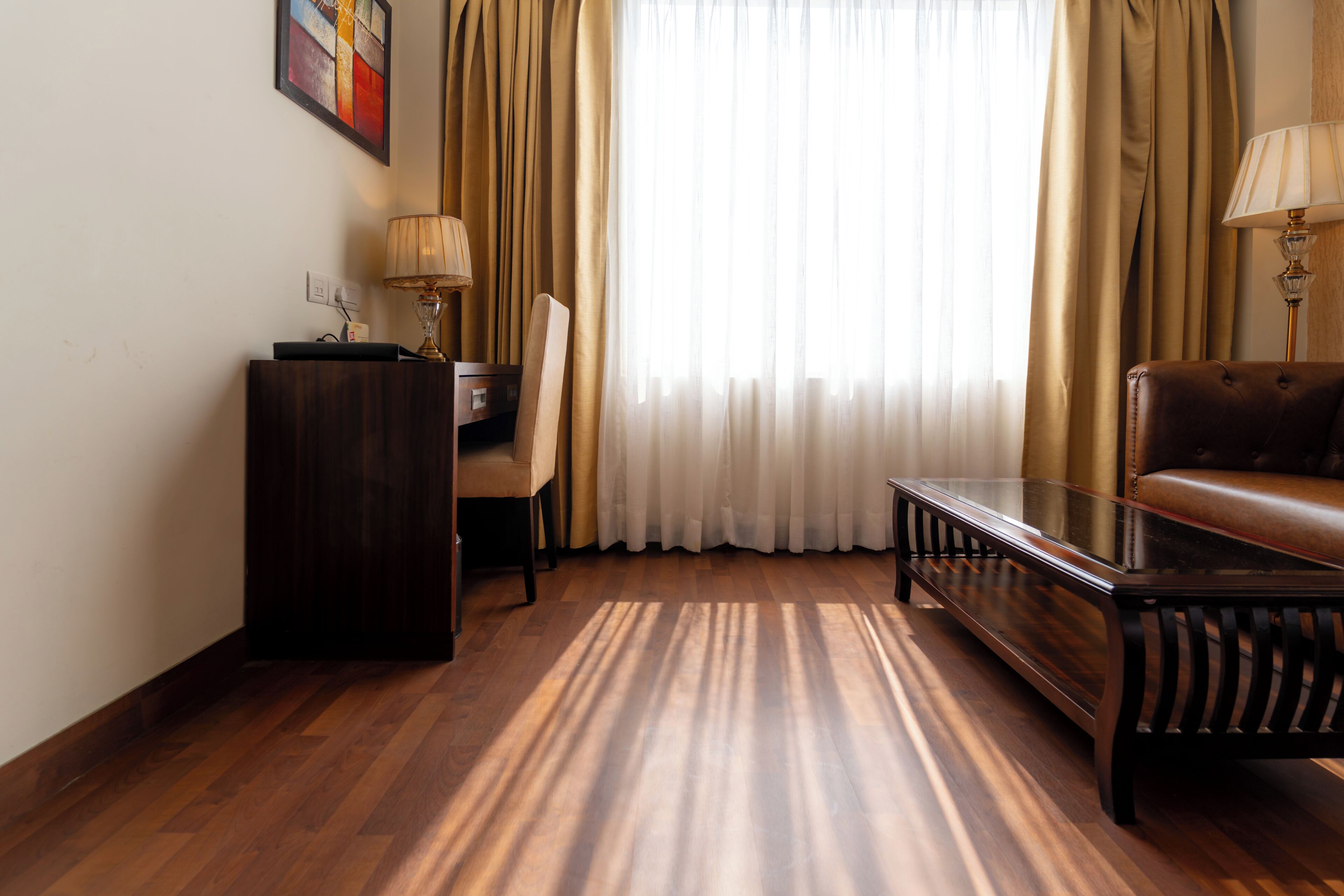 Een kamer met een houten vloer, een tafel met een glazen blad, een leren bank, een bureau met een lamp en een raam met gordijnen.