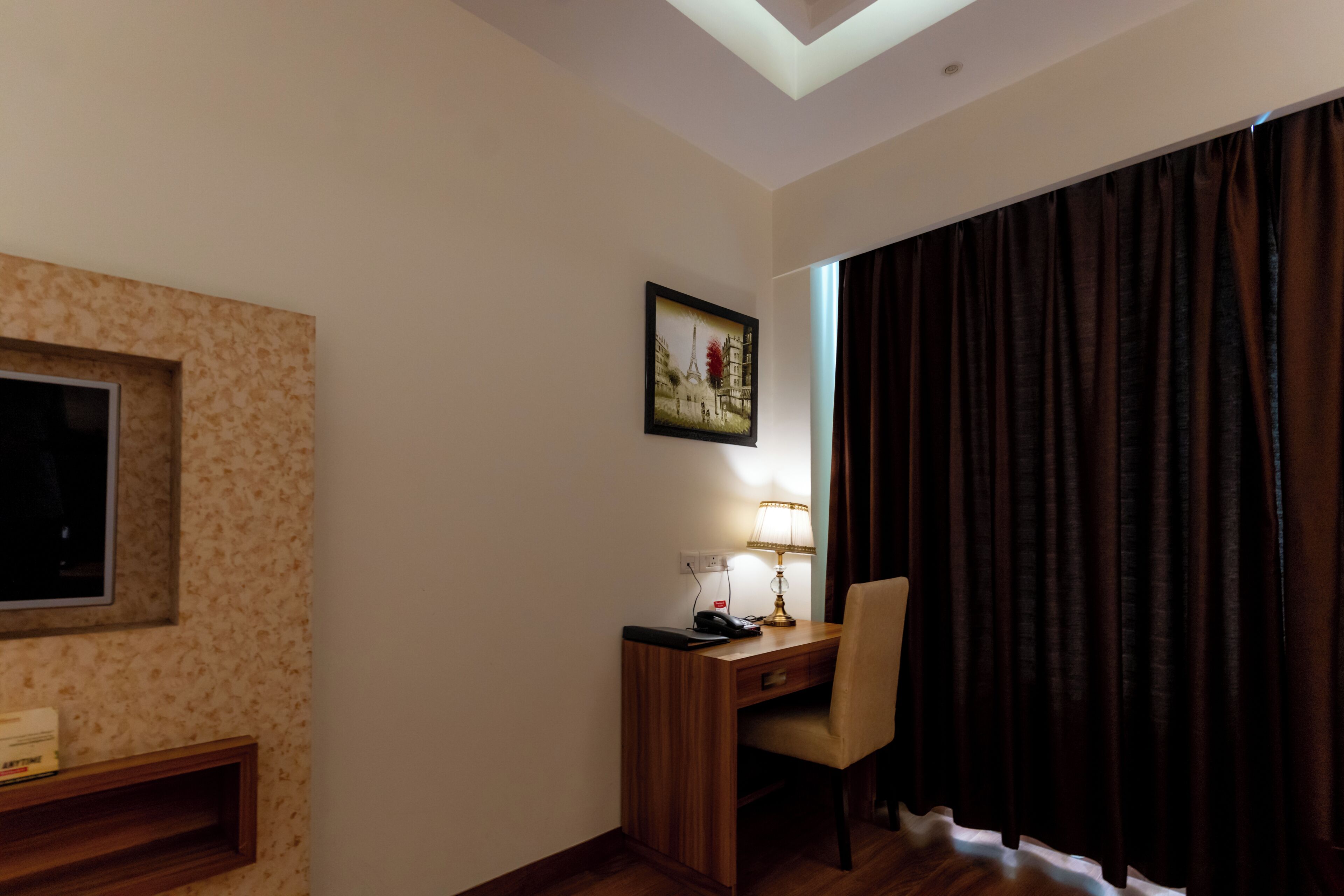 A hotel room with a desk, chair, lamp, picture of the Eiffel Tower, and a window with curtains.