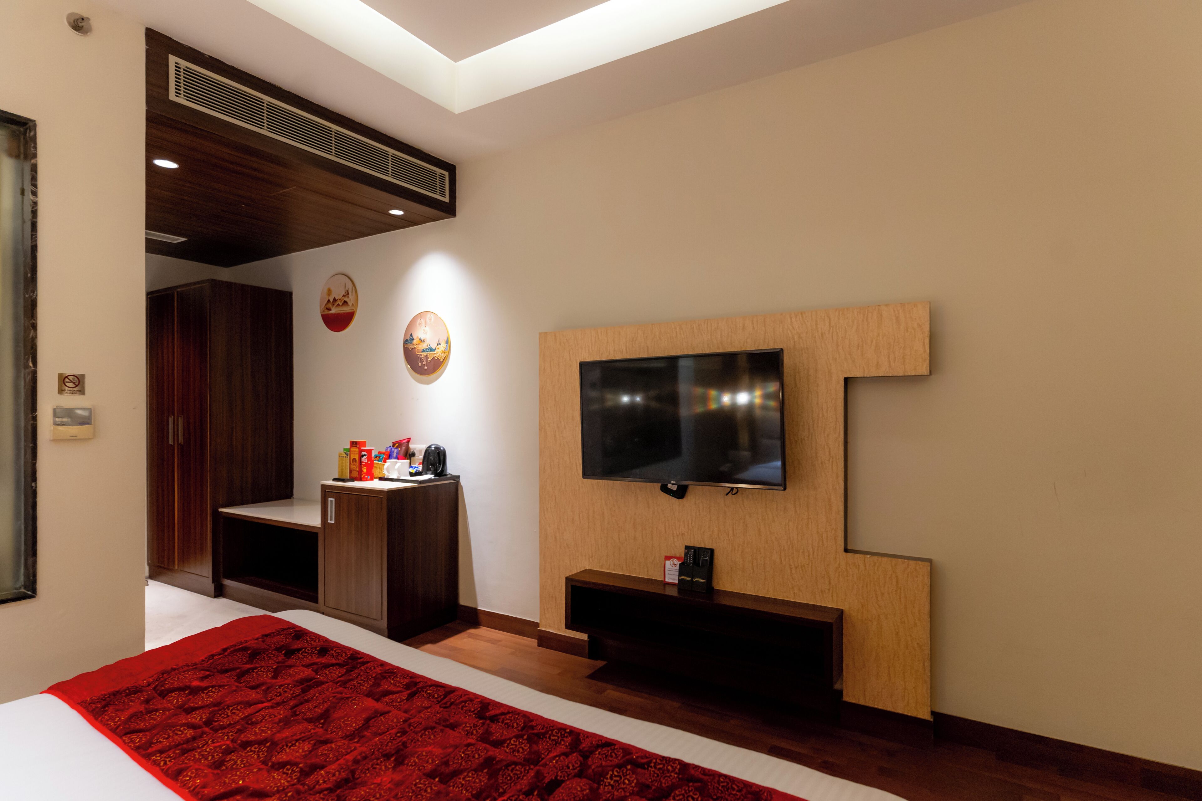 A modern hotel room with a large flat-screen TV mounted on a wooden panel, a built-in wardrobe, and a red patterned area rug.