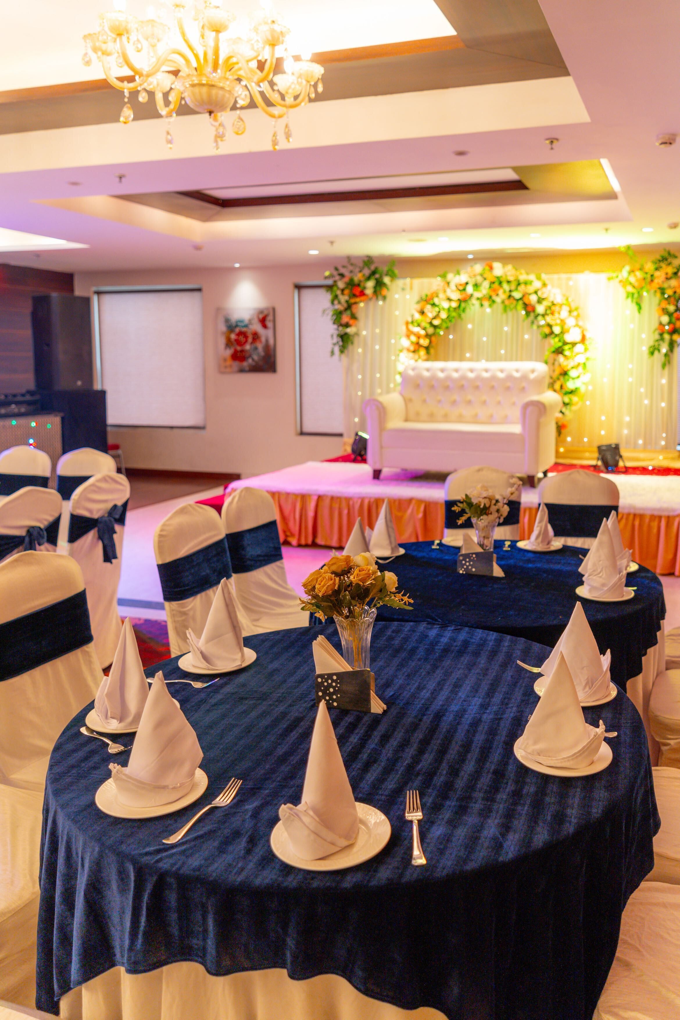 A banquet hall with round tables, chairs with blue sashes, and a stage with floral decorations.