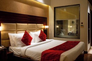 A hotel room with a large bed, a wooden headboard, a nightstand, a red throw blanket, and a large window with a view of the interior. - The Grand Ambassador (Phagwara)