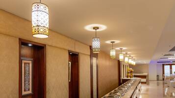 A long buffet table with multiple serving dishes, overhead patterned lighting, and wooden doors along the wall.