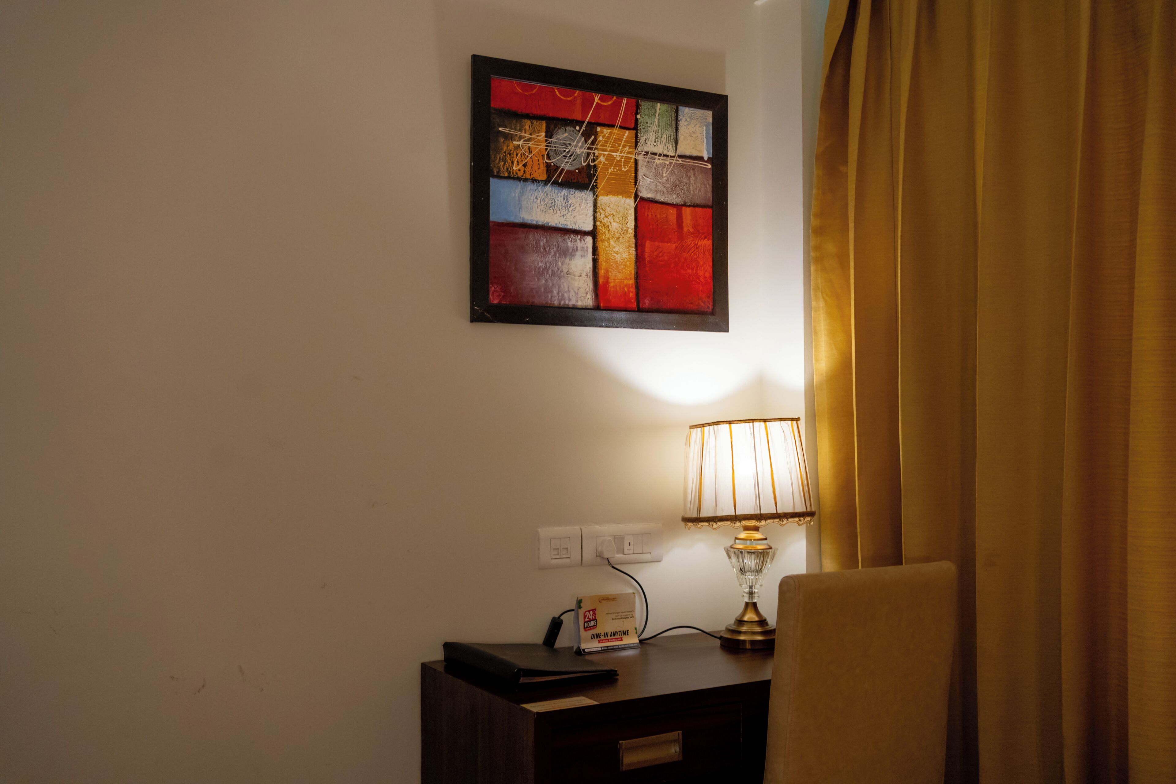 A room with a bedside table, a lamp, a power outlet, and a wall-mounted abstract painting.
