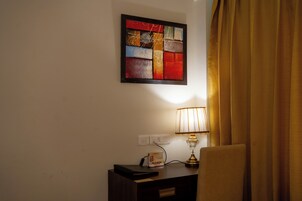 A room with a bedside table, a lamp, a power outlet, and a wall-mounted abstract painting.
