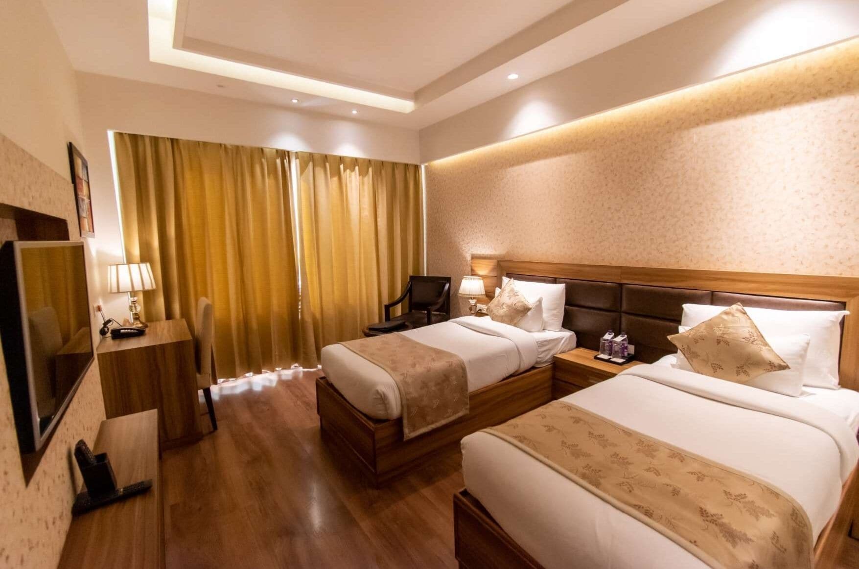 Habitación de hotel con dos camas, un escritorio, una silla, un televisor y un ventanal con cortinas.