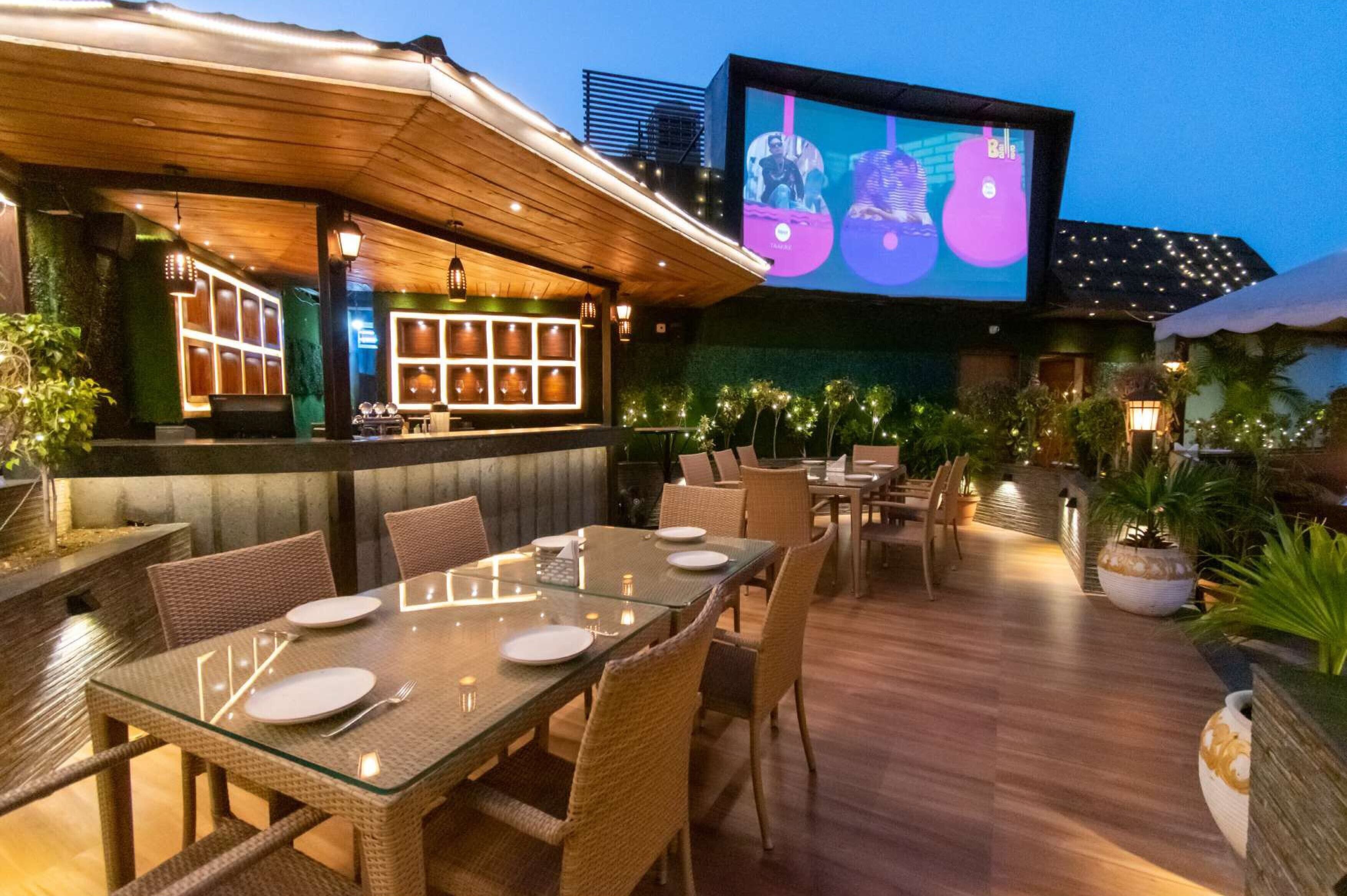 Un área de comedor al aire libre con mesas y sillas, un bar con una pantalla que muestra un video, y vistas del horizonte de la ciudad.