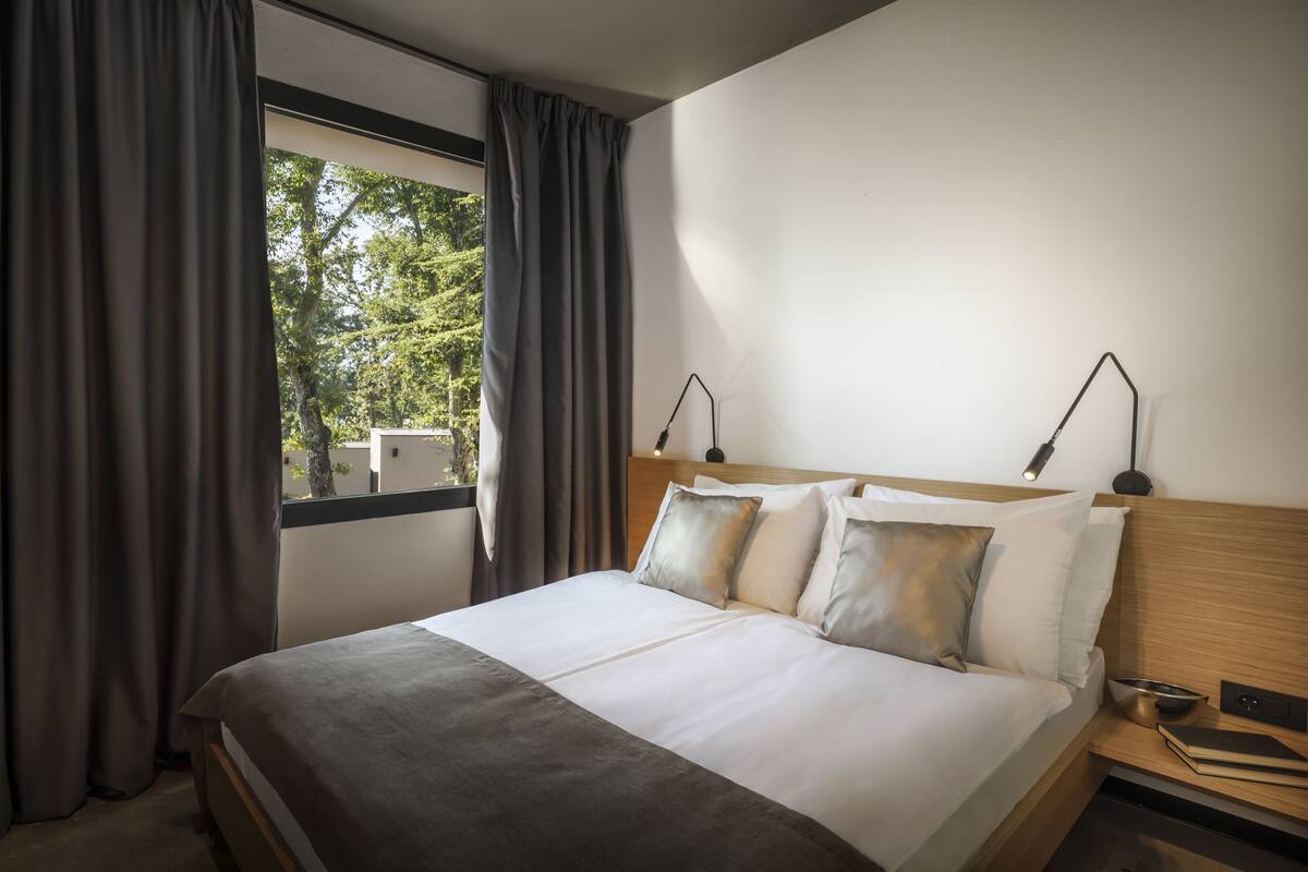 Una habitación de hotel con cama, dos lámparas de noche, mesita de noche, ventana con cortinas y vista a árboles.